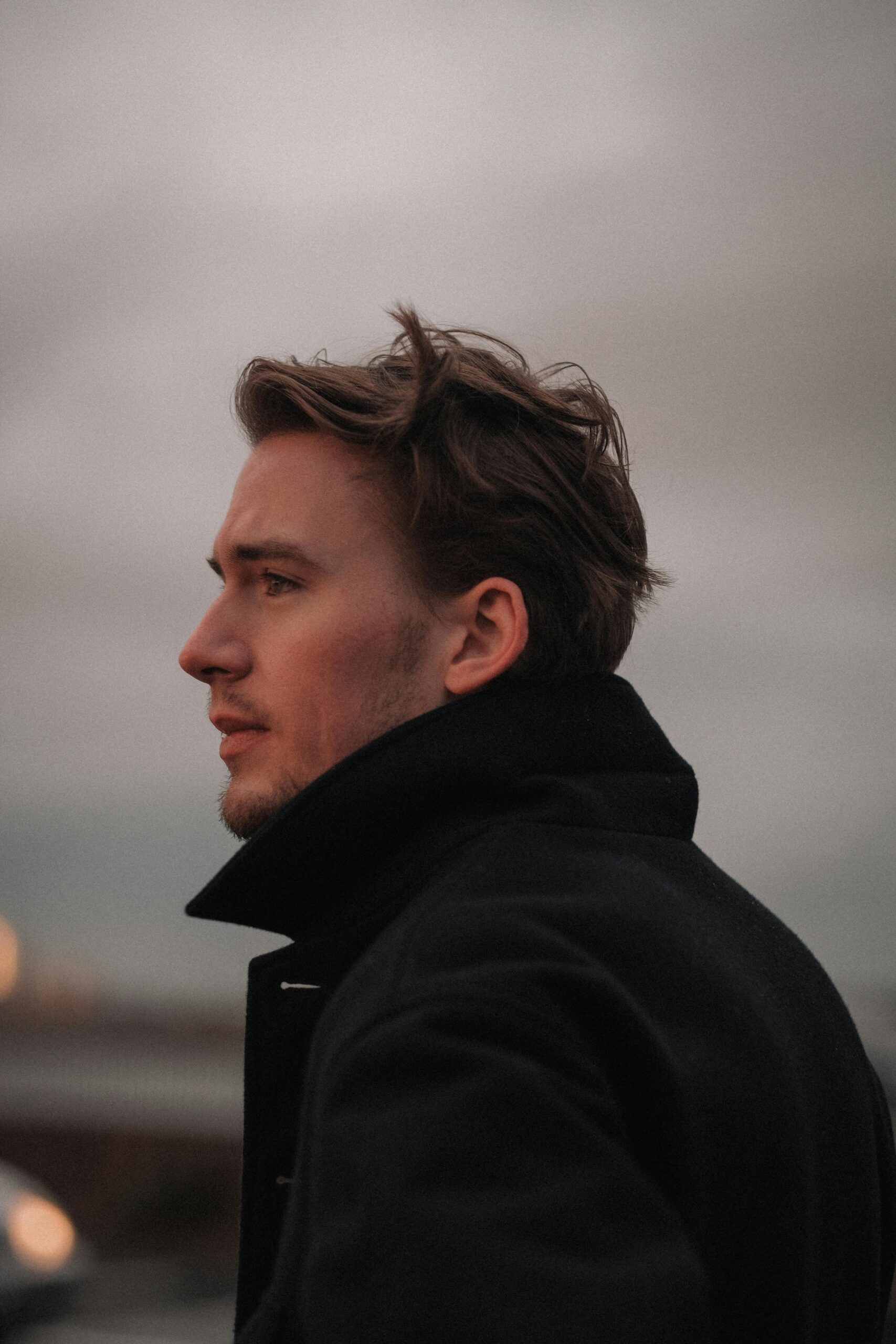 A thoughtful young man in profile wearing a dark coat, outdoors at sunset.