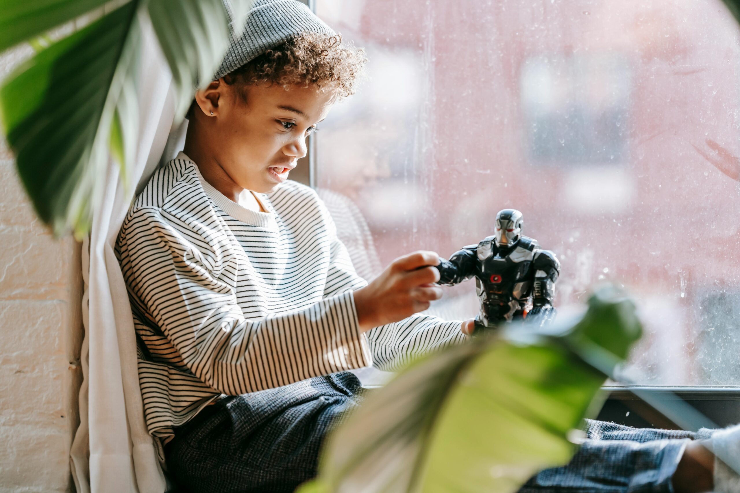 Side view of African American boy with curly hair sitting on windowsill and playing with toy at home in daytime