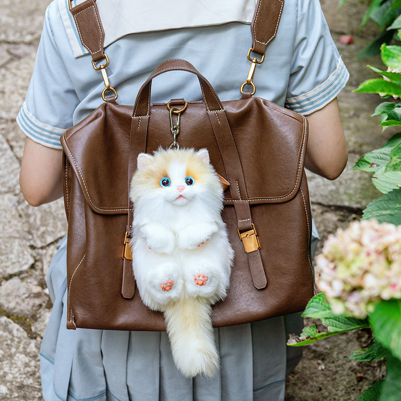 Back view of a realistic long-tailed orange cat plush charm hanging on a girl's brown leather backpack.