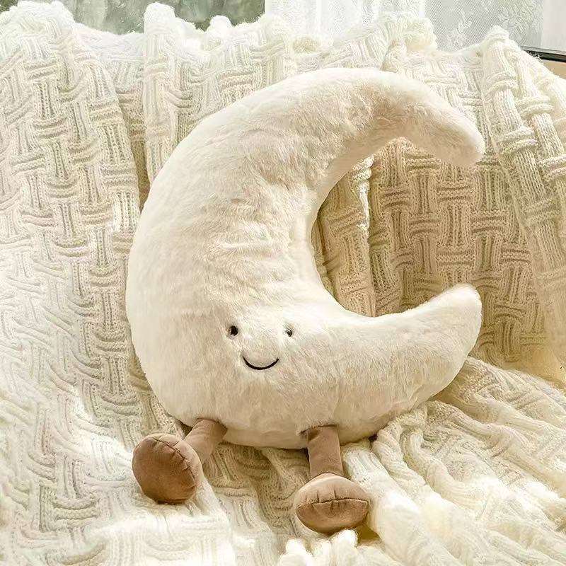 A white crescent moon plush pillow with a cute face sitting on a cozy white chair.